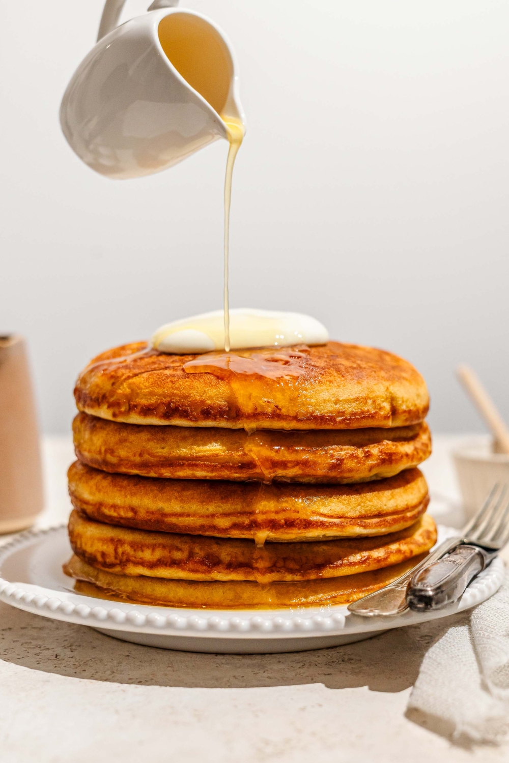 A white picture that has honey butter in it, dumping it on top of a stack of four honey butter, golden diner pancakes that are on a white plate.