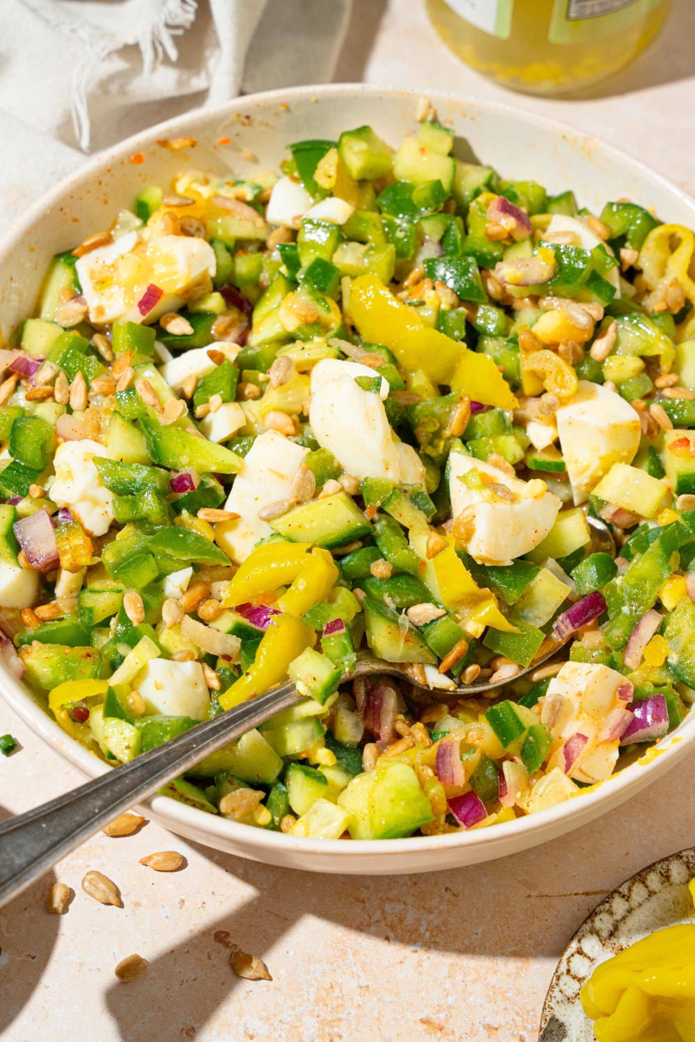 A white bowl that is filled with a hot girl summer salad consisting of chopped onion, green, bell pepper, cucumber, hard-boiled eggs, pepperoncini, and roasted sunflower kernels, all mixed in a zesty Italian dressing.