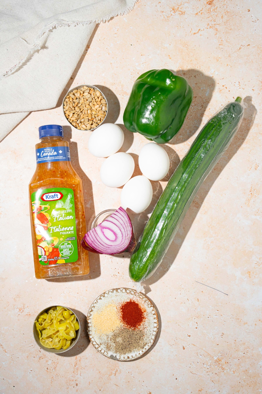 A bottle of zesty Italian dressing, a bowl of roasted sunflower seeds, a green, bell popper, four eggs, a zucchini, a half an onion, and a bowl of spices on the white counter.