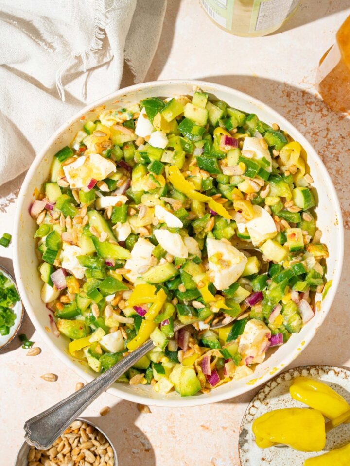 A hot girl summer salad consisting of chopped onion, green, bell pepper, cucumber, hard-boiled eggs, pepperoncini, and roasted sunflower kernels, all mixed in a zesty Italian dressing in a white bowl.