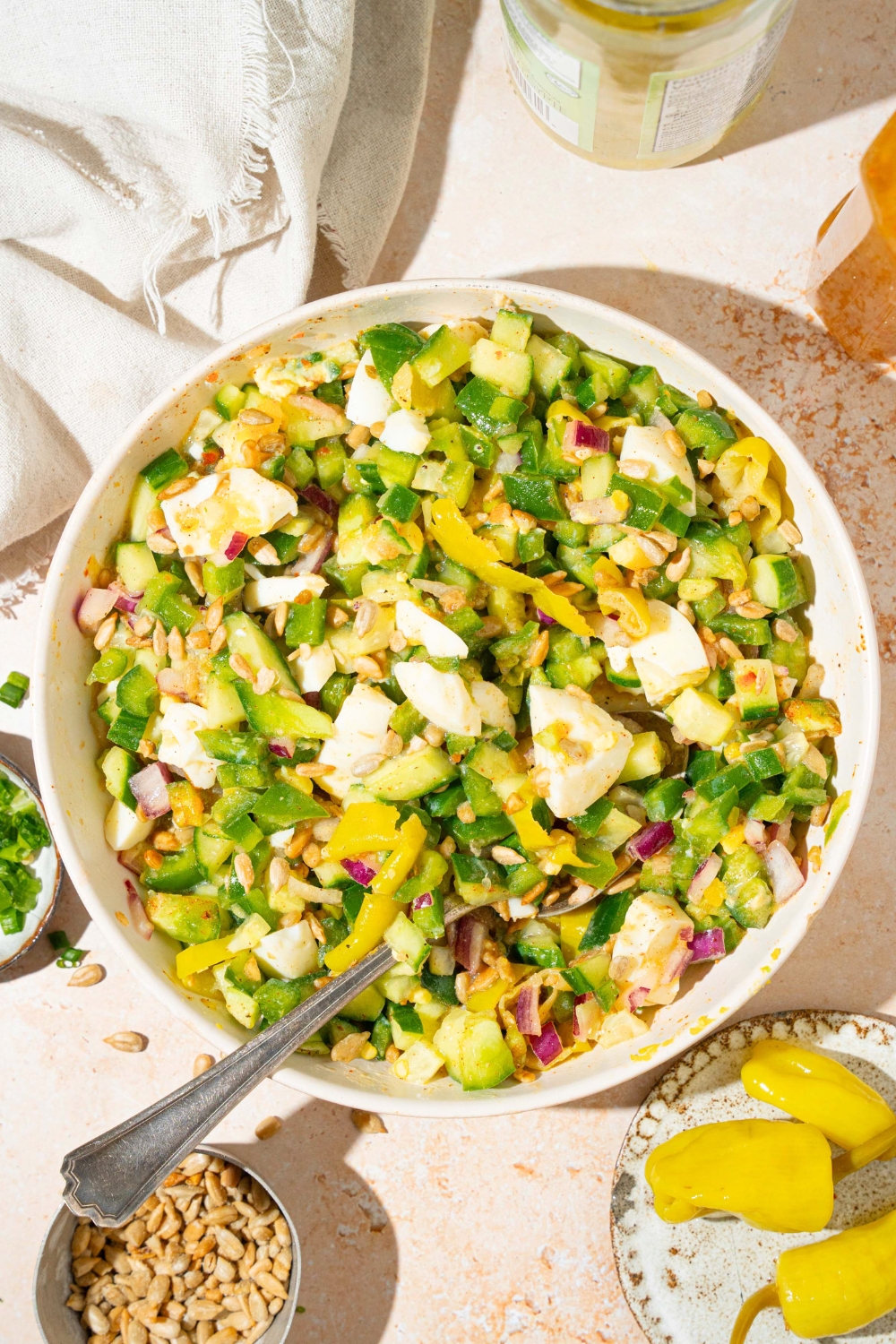 A hot girl summer salad consisting of chopped onion, green, bell pepper, cucumber, hard-boiled eggs, pepperoncini, and roasted sunflower kernels, all mixed in a zesty Italian dressing in a white bowl.