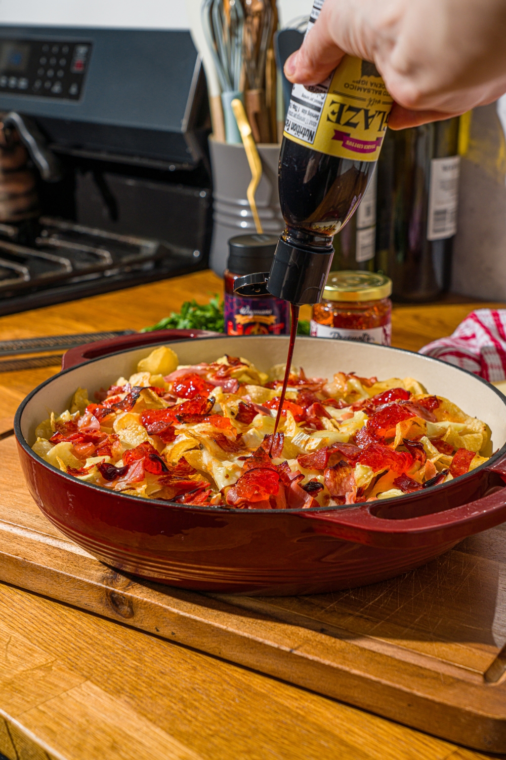 A baking dish with baked charcuterie nachos being drizzled with balsamic glaze. The dish is on a wooden board.