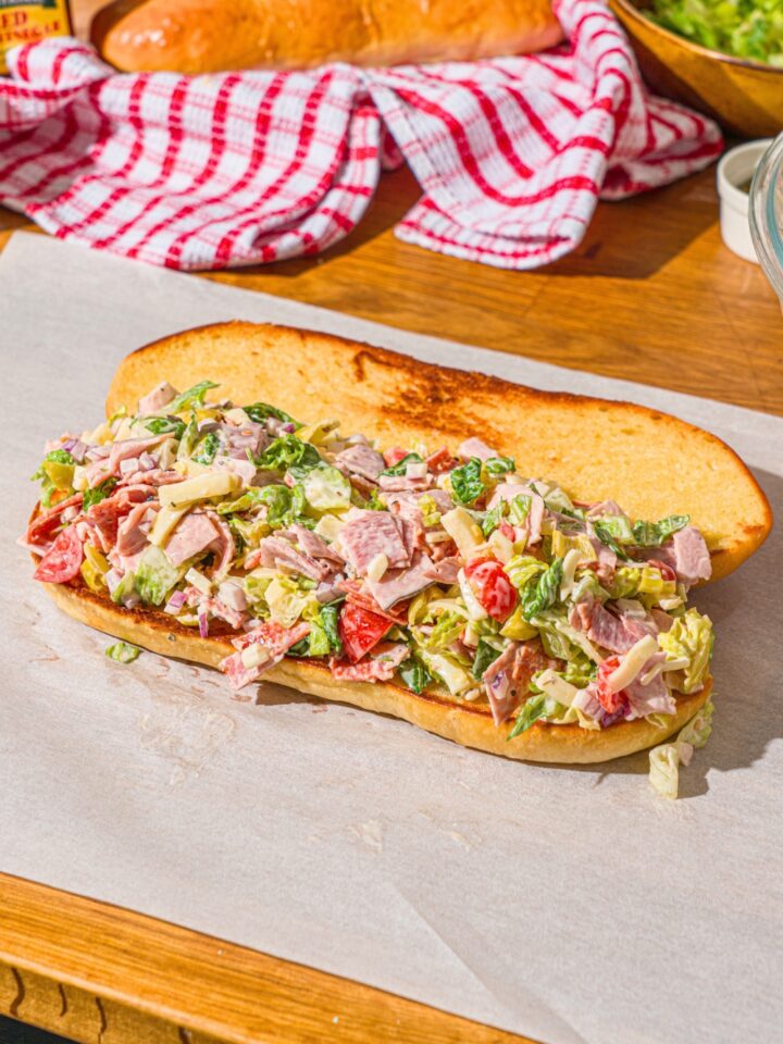 A piece of parchment paper with a whole open chopped Italian sandwich. The sandwich is on a wooden counter with a red striped napkin.