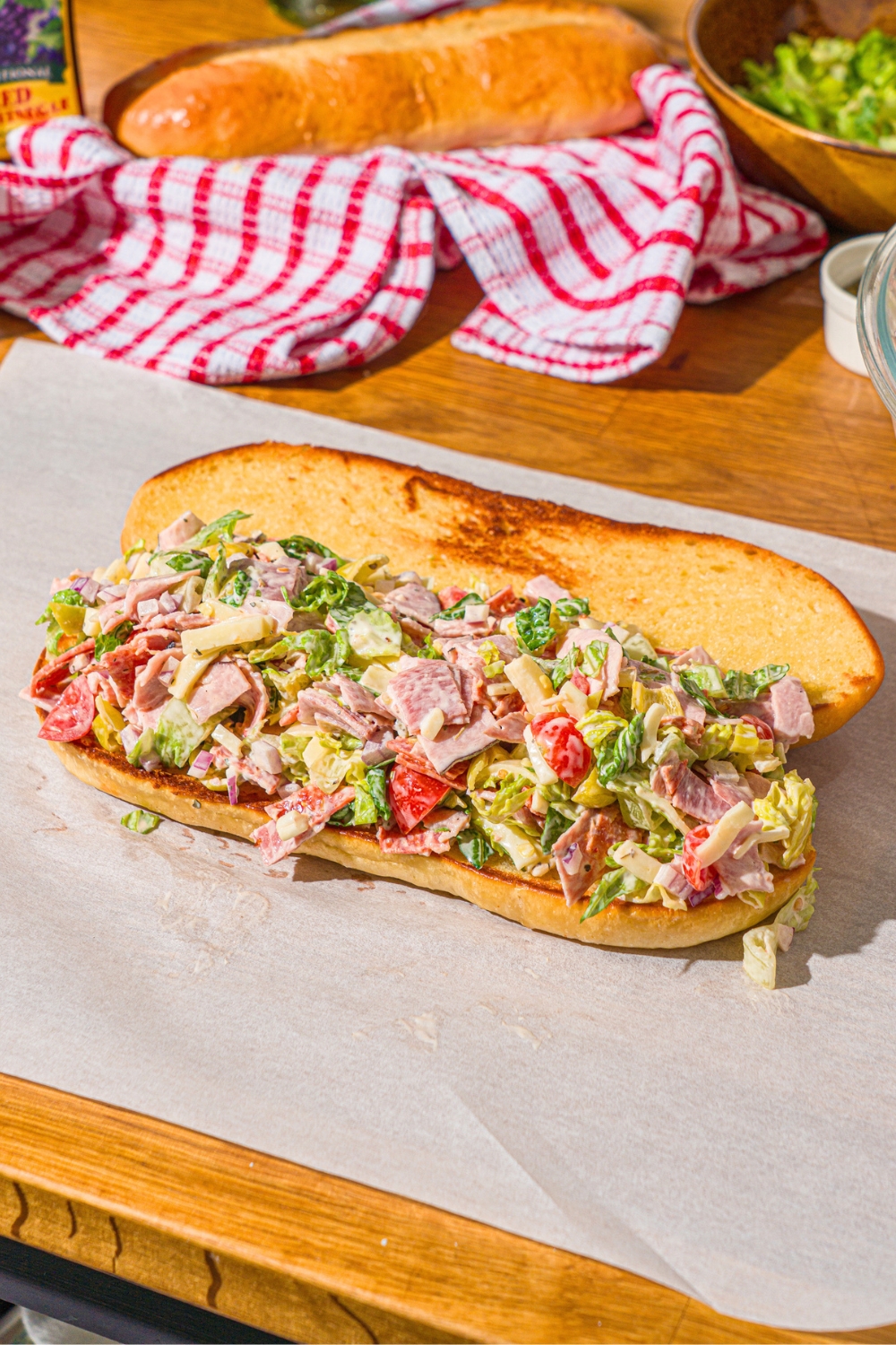 A piece of parchment paper with a whole open chopped Italian sandwich. The sandwich is on a wooden counter with a red striped napkin.