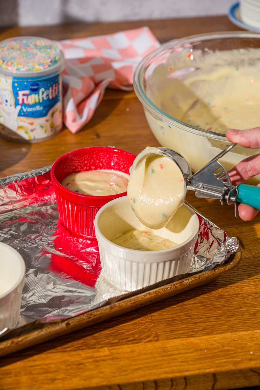 A hand scooping funfetti cake mix batter with a cookie scoop into a white ramekin. Behind that is a red ramekin filled with the cake batter and they're both on a foil lined baking sheet.