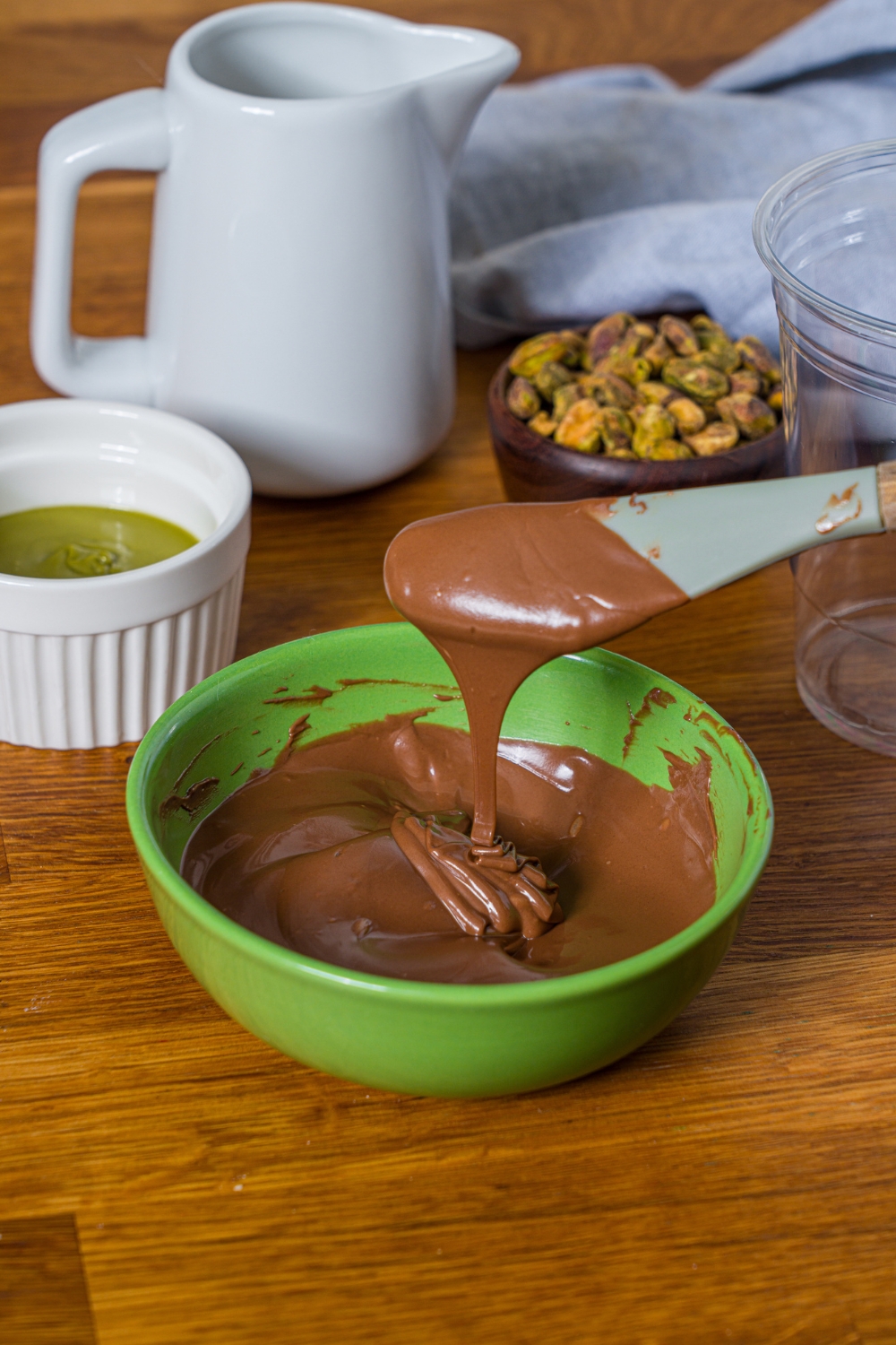 A rubber spatula with melted chocolate on it, drizzle it into a green bowl that is filled with the belted chocolate on top of the wooden counter.