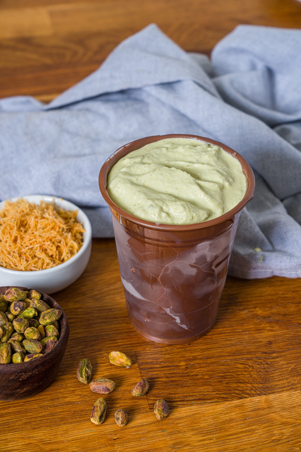 A Dubai pistachio shake in a chocolate lined cup on a wooden counter. Next to it is a bowl of pistachios in a bowl of katafi.