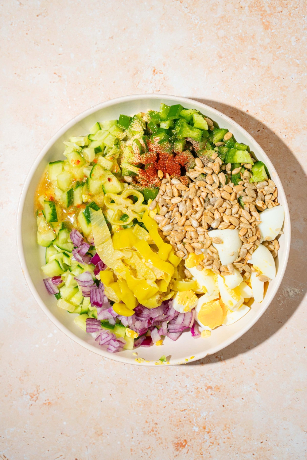 Sunflower kernels, chopped, hard-boiled eggs, chopped pepperocini, chopped onion, and chopped cucumber all in a white bowl.
