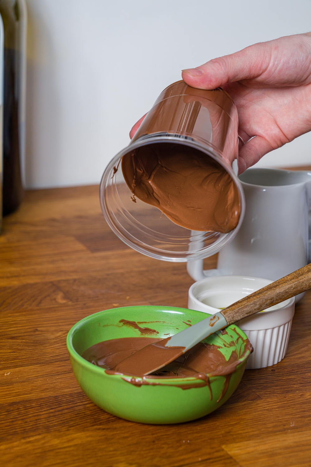A hand holding a clear plastic cup that is lined with melted chocolate over a green bowl that is filled with the melted chocolate.