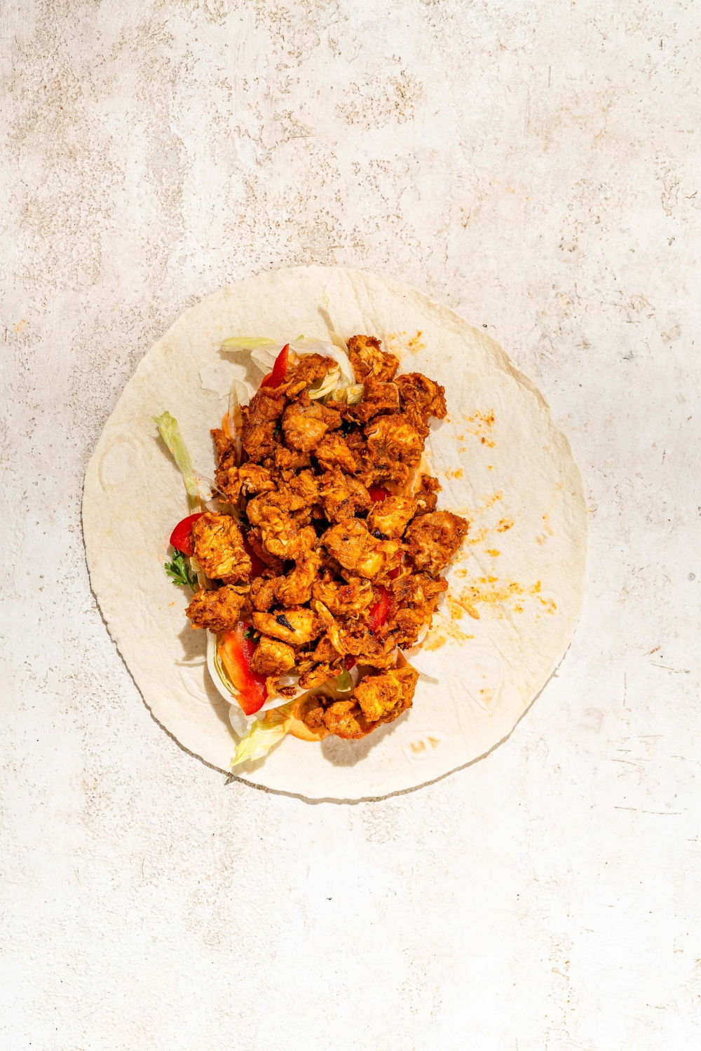 An open tortilla wrap with chipotle mayo, red pepper, shredded lettuce, and chipotle chicken. The wrap is on a tan counter.