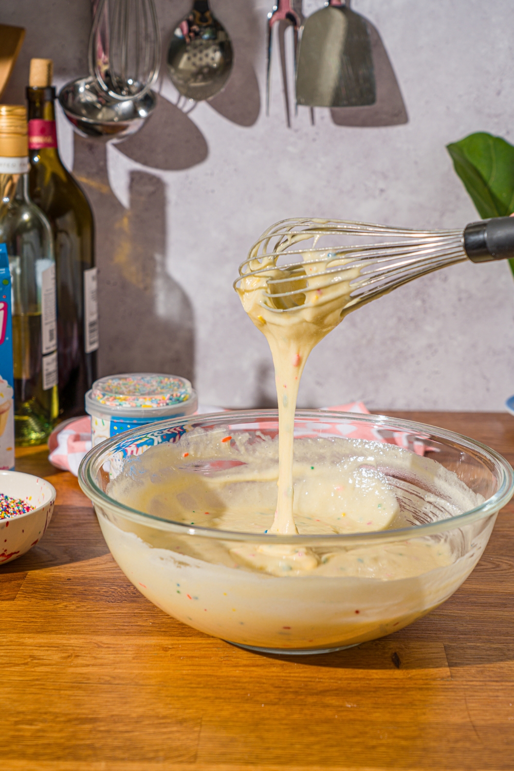 A whisk that has funfetti mix batter on it drizzling it into a glass bowl that is filled with the cake batter.