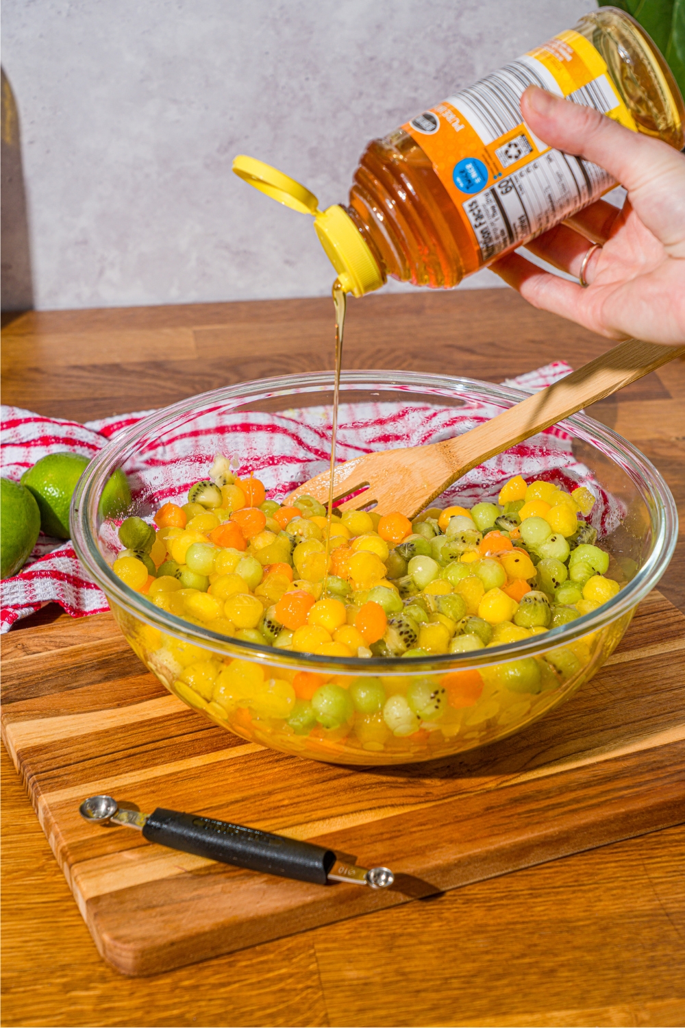 A hand squeezing honey into a large glass bowl that is filled with dot fruit salad.
