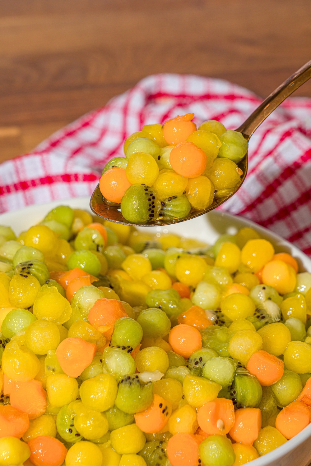 A spoon with balls of kiwi, melon, yellow watermelon, and apricots on it over a bowl of the dot fruit salad.