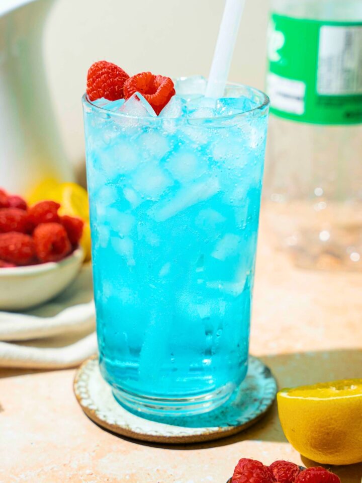 A tall glass that is filled with pebble ice and a copycat Sonic ocean water drink. There are some raspberries on top of the drink and a straw is in it.
