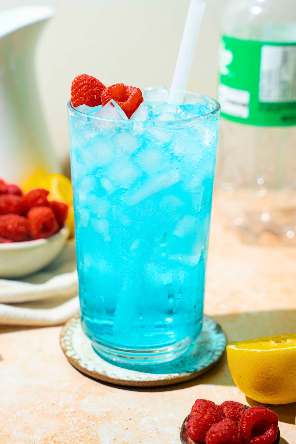 A tall glass that is filled with pebble ice and a copycat Sonic ocean water drink. There are some raspberries on top of the drink and a straw is in it.