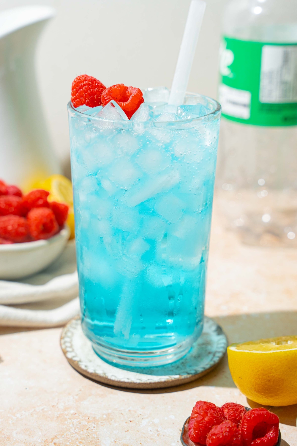 A tall glass of ocean water drink served with pebble ice, a straw, and garnished with fresh raspberries. The glass is on a coaster on a tan counter with a bottle and can of soda, bowls of raspberries, and sliced lemon.
