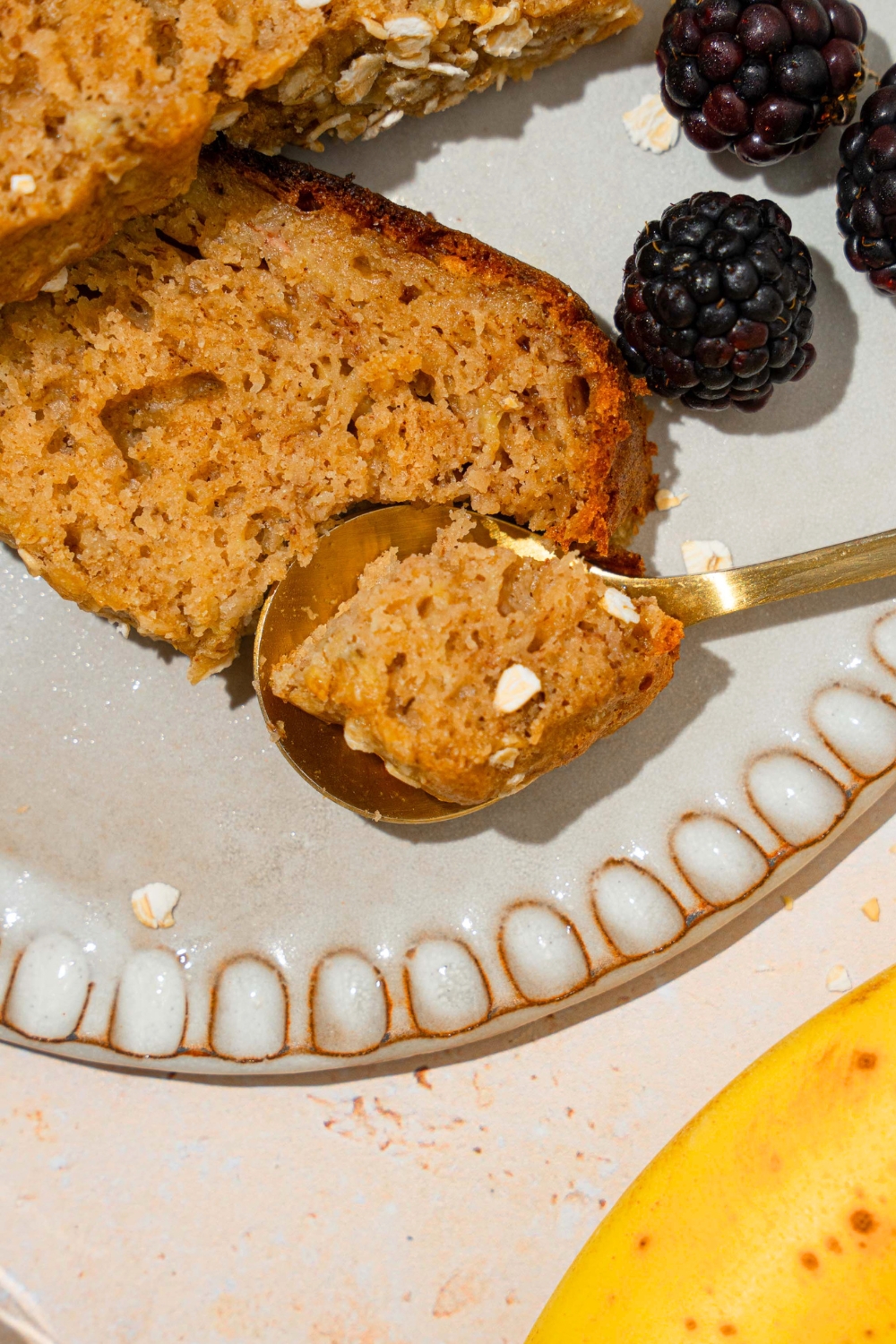 A piece of rice cooker, banana bread on top of a gold spoon that is on a white plate. Behind the spoon is a slice of the banana bread that the piece is taken from.