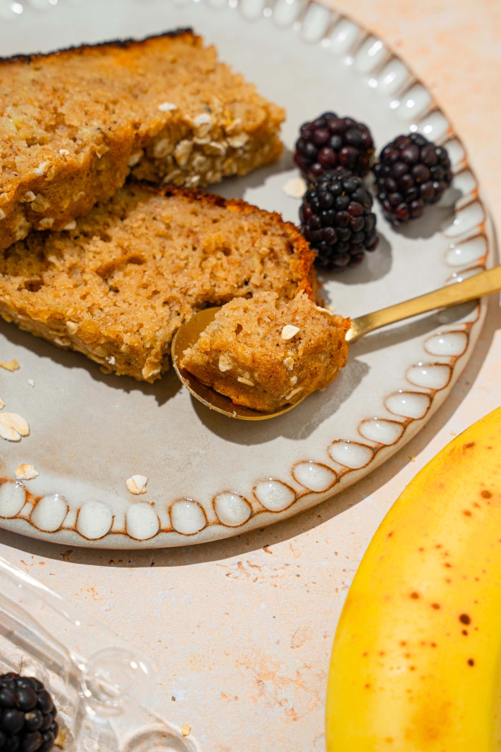 A piece of rice cooker, banana bread on a gold spoon with two slices of the banana bread behind it on a white plate.