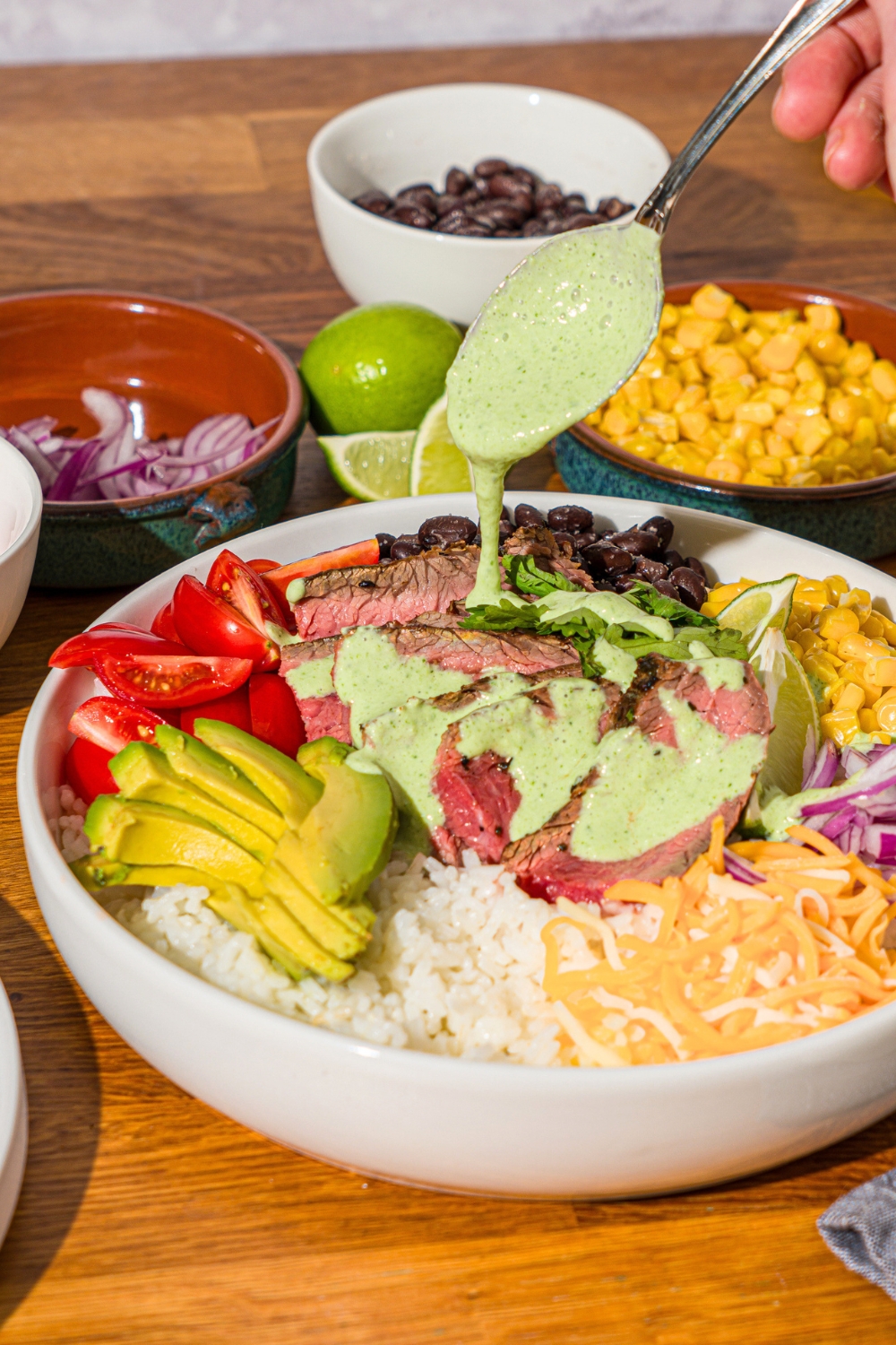 A spoon drizzling a cilantro lime crema on top of a steak bowl that has sliced steak, avocado, peppers, onions, corn, shredded cheese, and white rice.