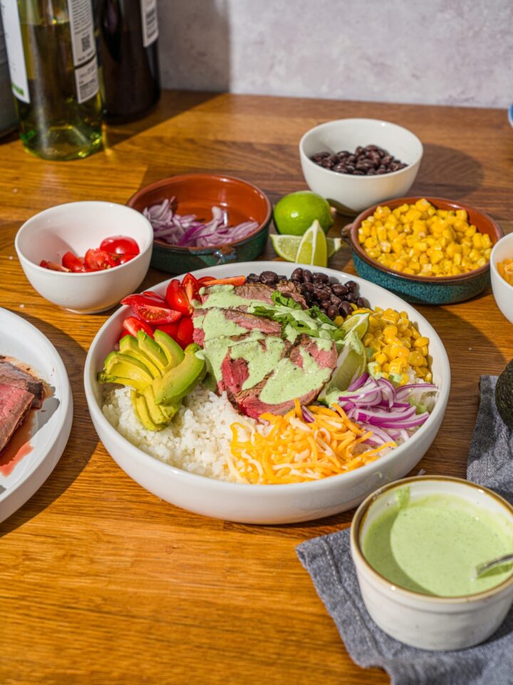 A large white bowl with a steak bowl served with rice, grilled marinated steak, corn, avocado, beans, tomato, red onion, and shredded cheese drizzled with a cilantro lime sauce. The bowl is on a wooden counter with several bowls of toppings.