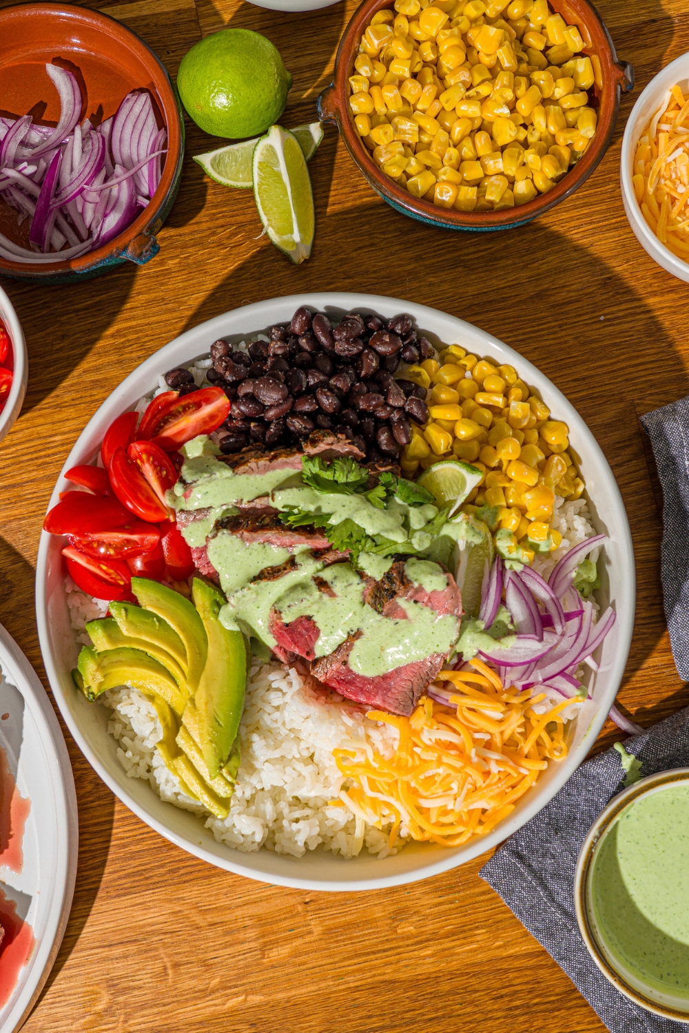 A large white bowl with a steak bowl served with rice, grilled marinated steak, corn, avocado, beans, tomato, red onion, and shredded cheese drizzled with a cilantro lime sauce. The bowl is on a wooden counter with several bowls of toppings.