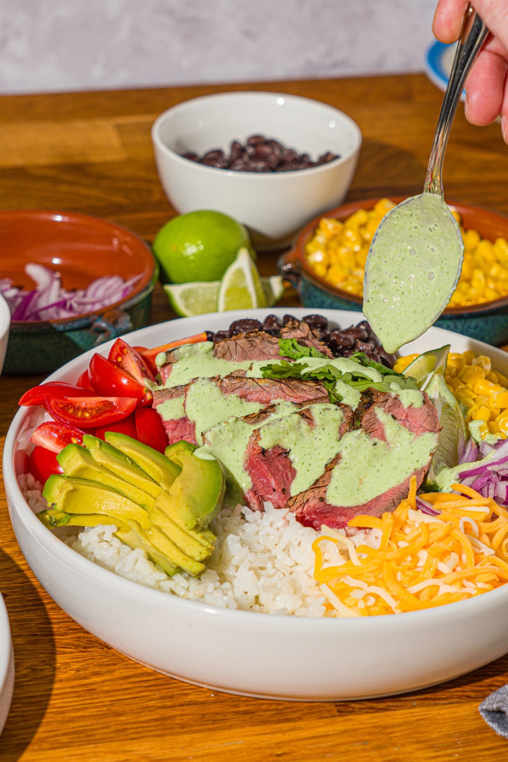 A large white bowl with a steak bowl served with rice, grilled marinated steak, corn, avocado, beans, tomato, red onion, and shredded cheese A spoon is drizzling cilantro lime sauce over the bowl. The bowl is on a wooden counter with several bowls of toppings.