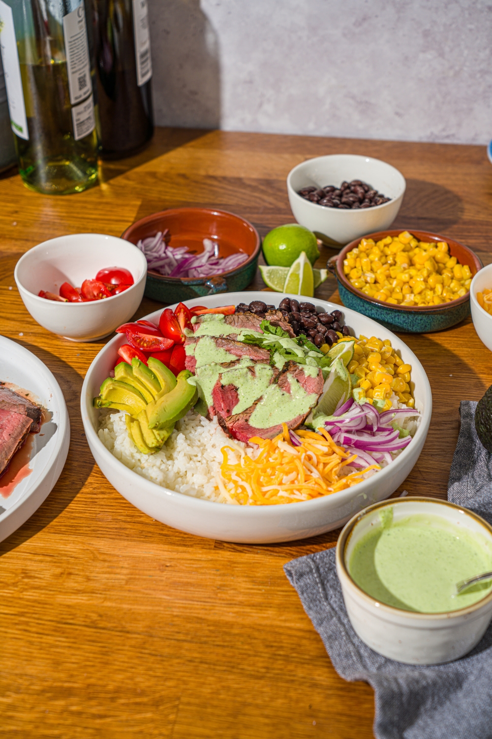 A large white bowl with a steak bowl served with rice, grilled marinated steak, corn, avocado, beans, tomato, red onion, and shredded cheese drizzled with a cilantro lime sauce. The bowl is on a wooden counter with several bowls of toppings.