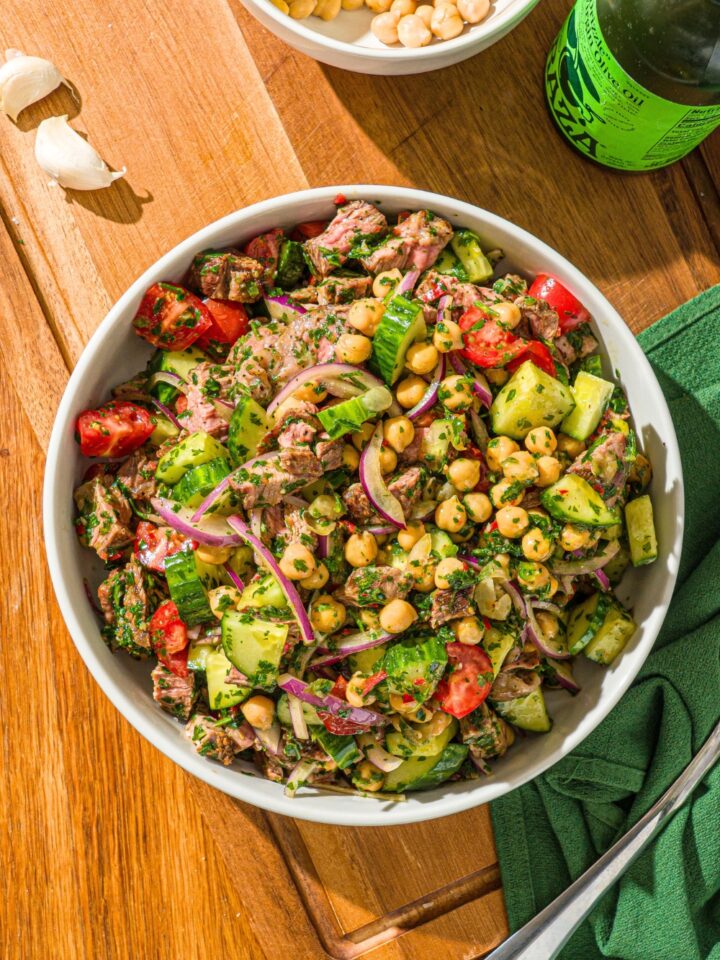 A steak salad with steak pieces, cucumber, chickpeas, and sliced red onion, all tossed in a chimichurri sauce in a white bowl on top of a wooden counter.