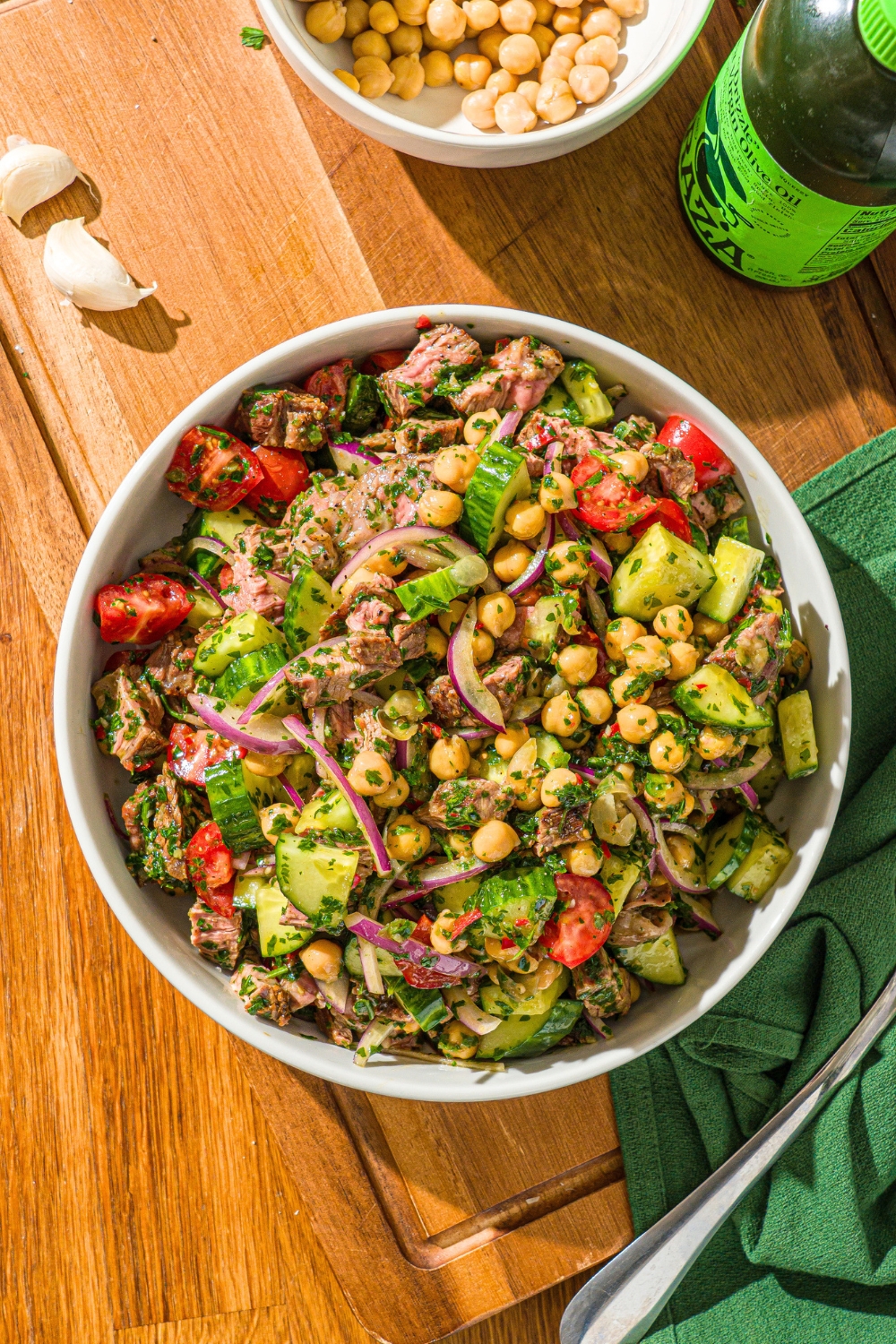 A steak salad with steak pieces, cucumber, chickpeas, and sliced red onion, all tossed in a chimichurri sauce in a white bowl on top of a wooden counter.
