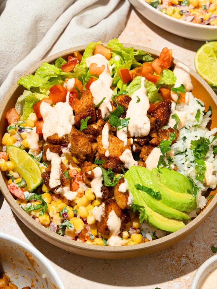 A street corn chicken rice bowl in a bowl. The bowl has seasoned seared chicken, avocado, street corn, lettuce, tomatoes, a lime wedge, and avocado with a cilantro lime dressing drizzled on top.