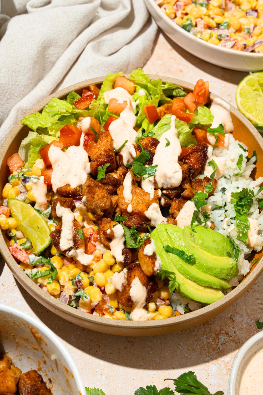 A street corn chicken rice bowl in a bowl. The bowl has seasoned seared chicken, avocado, street corn, lettuce, tomatoes, a lime wedge, and avocado with a cilantro lime dressing drizzled on top.