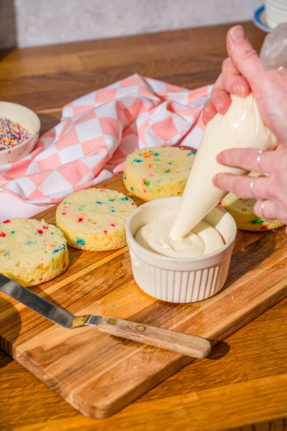 Two hands holding a bag of vanilla frosting, piping it into a white ramekin that has a dot cake in it. Behind it are three more circular dot cakes.
