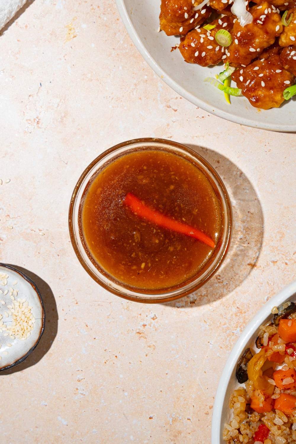A small bowl with general tso sauce with a red chile. The bowl is on a tan counter with a plate of general tso's chicken.