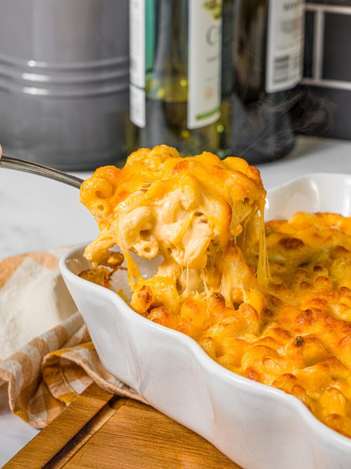 A baking pan with baked bacon macaroni and cheese with a spoon taking a scoop from the pan. The pan is on a wooden board with a yellow checkered napkin.