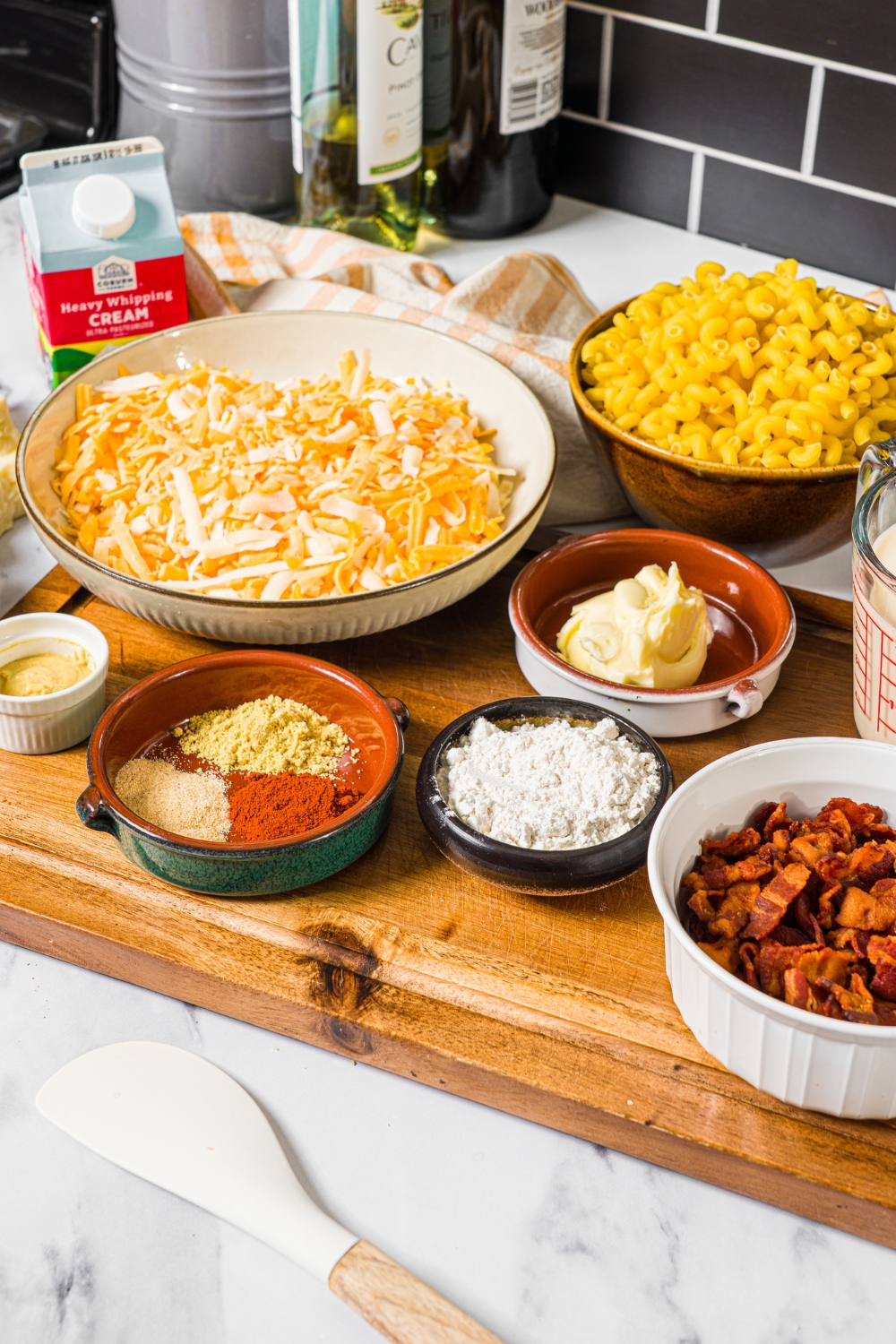 A wooden board with several bowls containing ingredients to make bacon mac and cheese including crispy bacon, butter, flour, cream, shredded cheeses, macaroni, and seasonings.