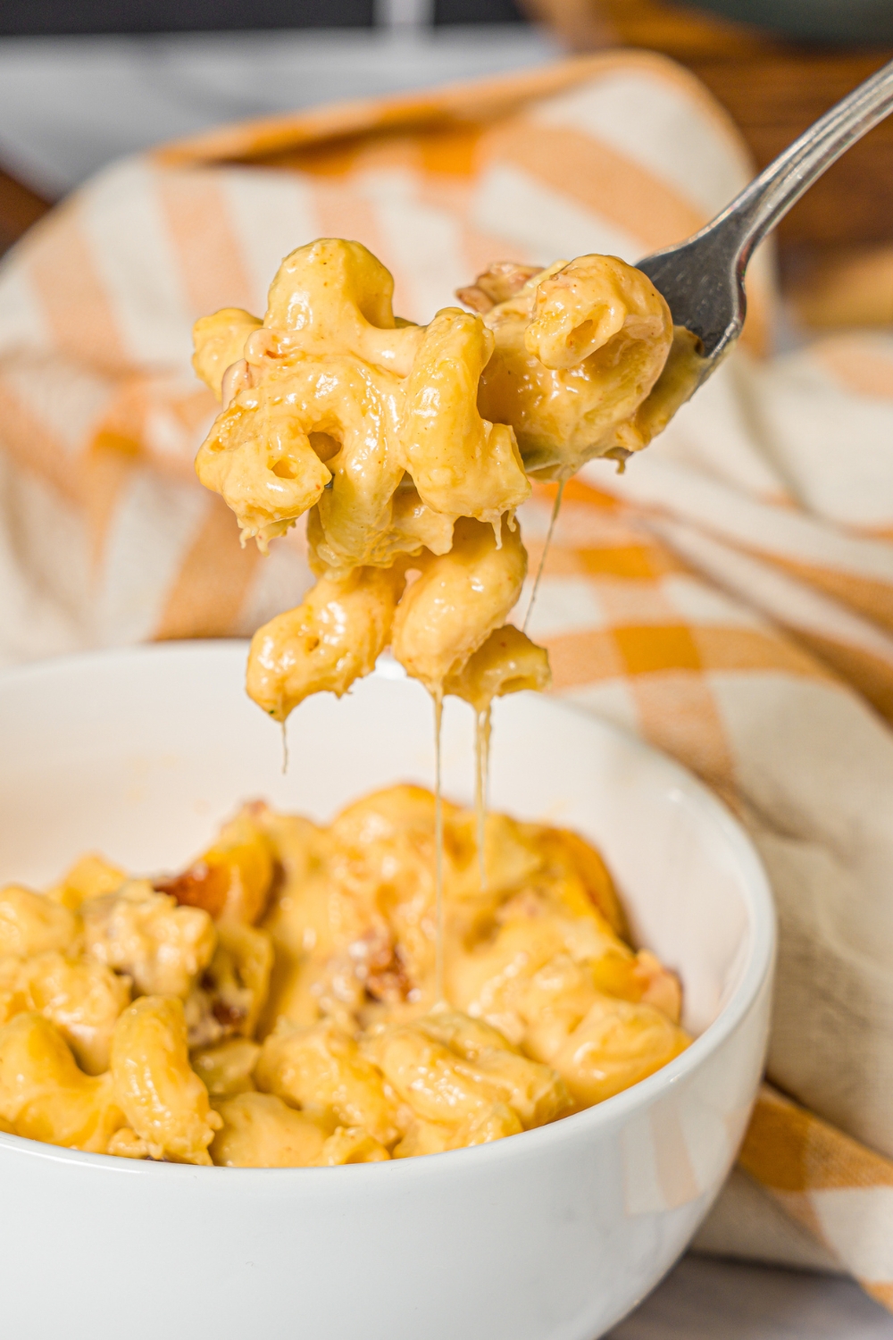 A close up of a fork with a bite of bacon macaroni and cheese. There is a bowl of mac and cheese and a yellow checkered napkin blurred in the background.