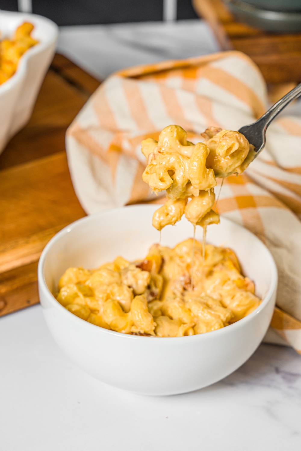 A bowl of bacon macaroni and cheese with a fork taking a bite from the bowl. The bowl is on a marbled counter with a yellow checkered napkin.