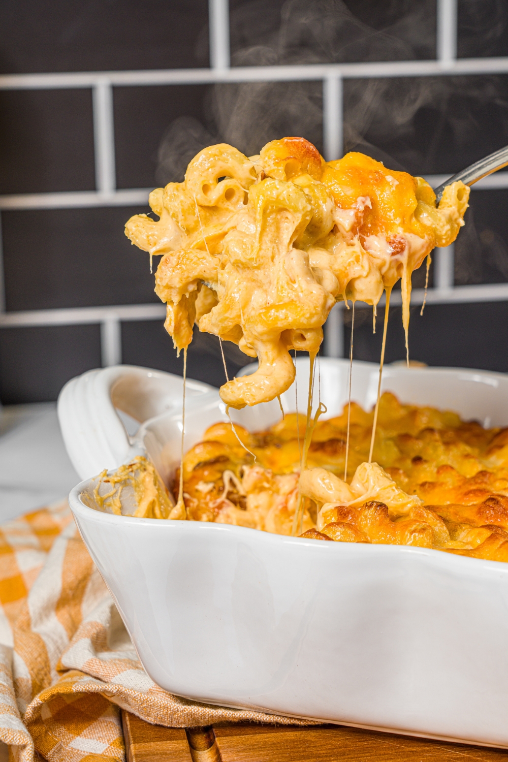 A baking pan with baked bacon macaroni and cheese with a spoon taking a scoop from the pan. The pan is on a wooden board with a yellow checkered napkin.
