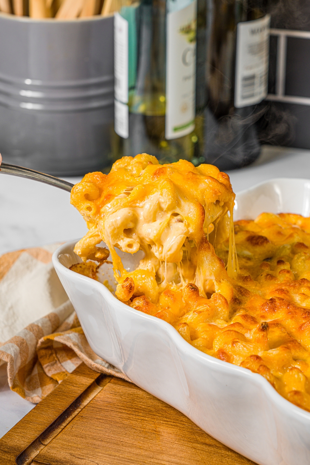 A baking pan with baked bacon macaroni and cheese with a spoon taking a scoop from the pan. The pan is on a wooden board with a yellow checkered napkin.
