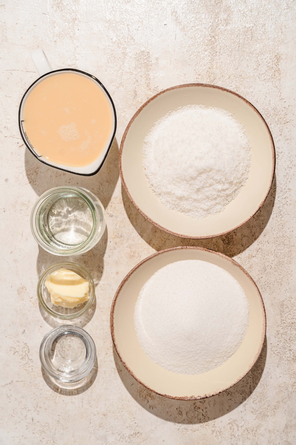 A tan counter with several bowls containing ingredients to make bala baiana including butter, condensed milk, shredded coconut, sugar, water, and vinegar.