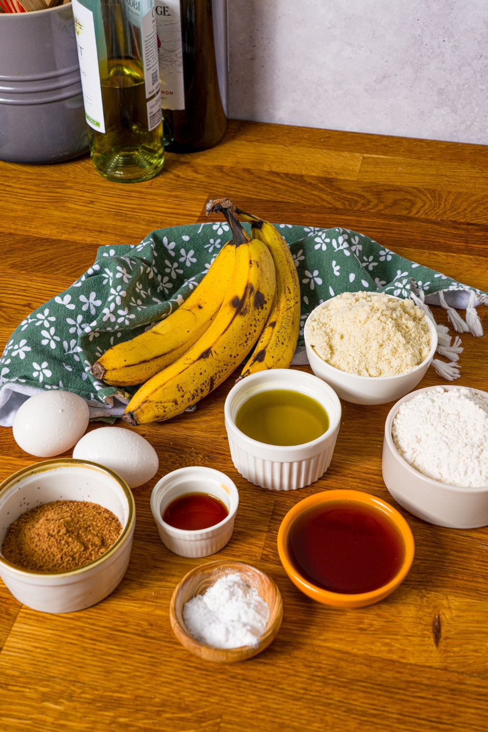 A wooden counter with bowls containing ingredients to make banana bread with olive oil including bananas, olive oil, egg, maple syrup, vanilla extract, coconut sugar, almond flour, cinnamon, and more.
