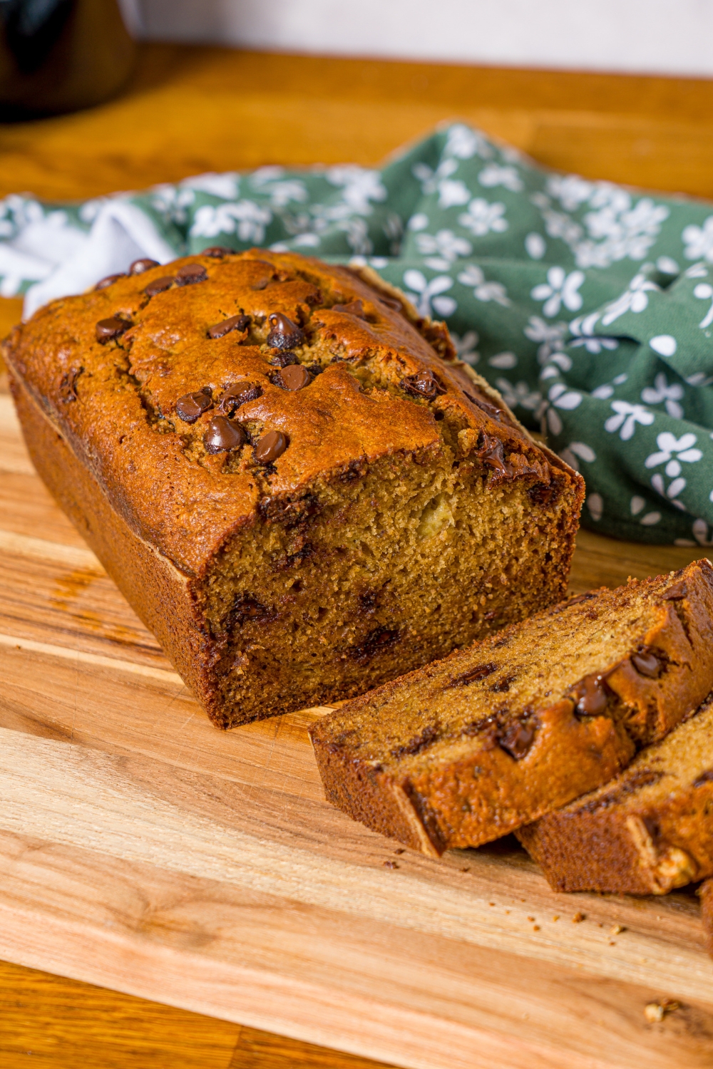 A wooden board with a loaf of chocolate chip banana bread with olive oil. Several slices have been sliced from the loaf. The board is on a wooden counter with a green floral napkin.