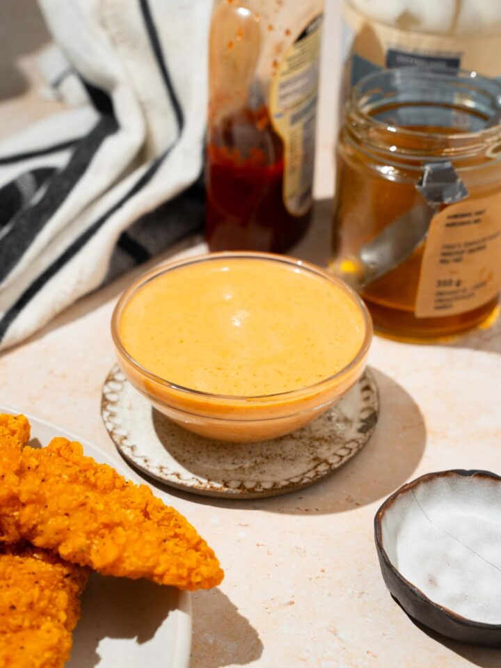 A bowl of bang bang sauce on a saucer on a tan counter with a plate of chicken tenders, jar of honey, and black striped napkin.
