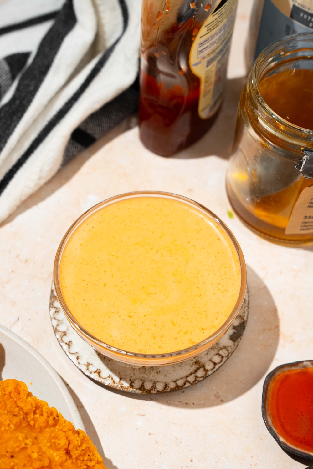 A bowl of bang bang sauce on a saucer on a tan counter with honey and a black striped napkin.