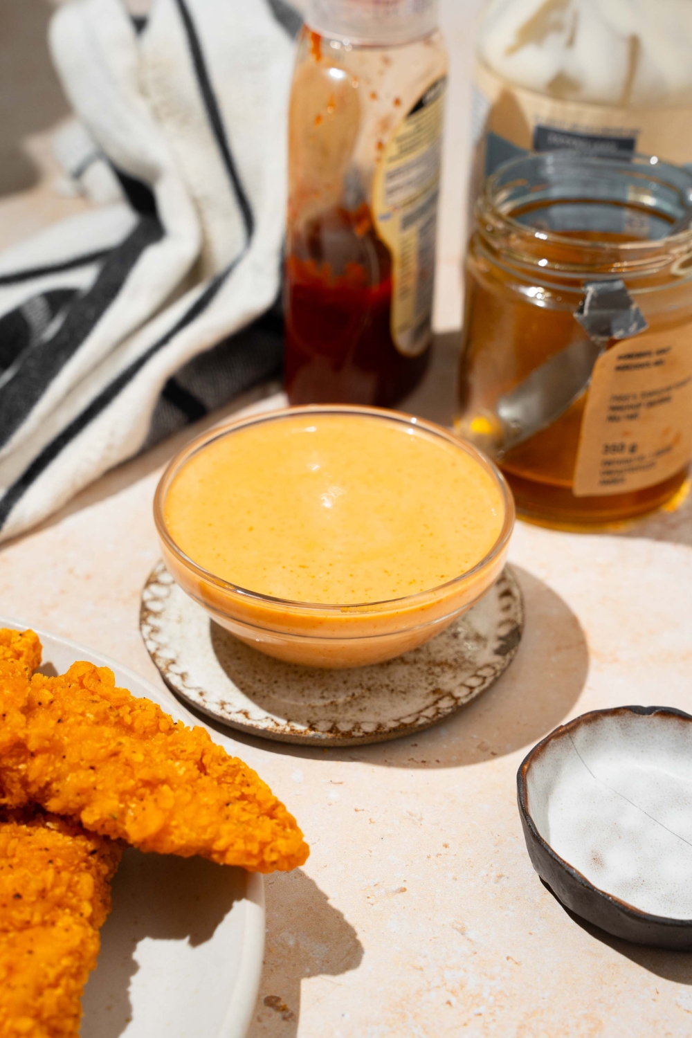 A bowl of bang bang sauce on a saucer on a tan counter with a plate of chicken tenders, jar of honey, and black striped napkin.