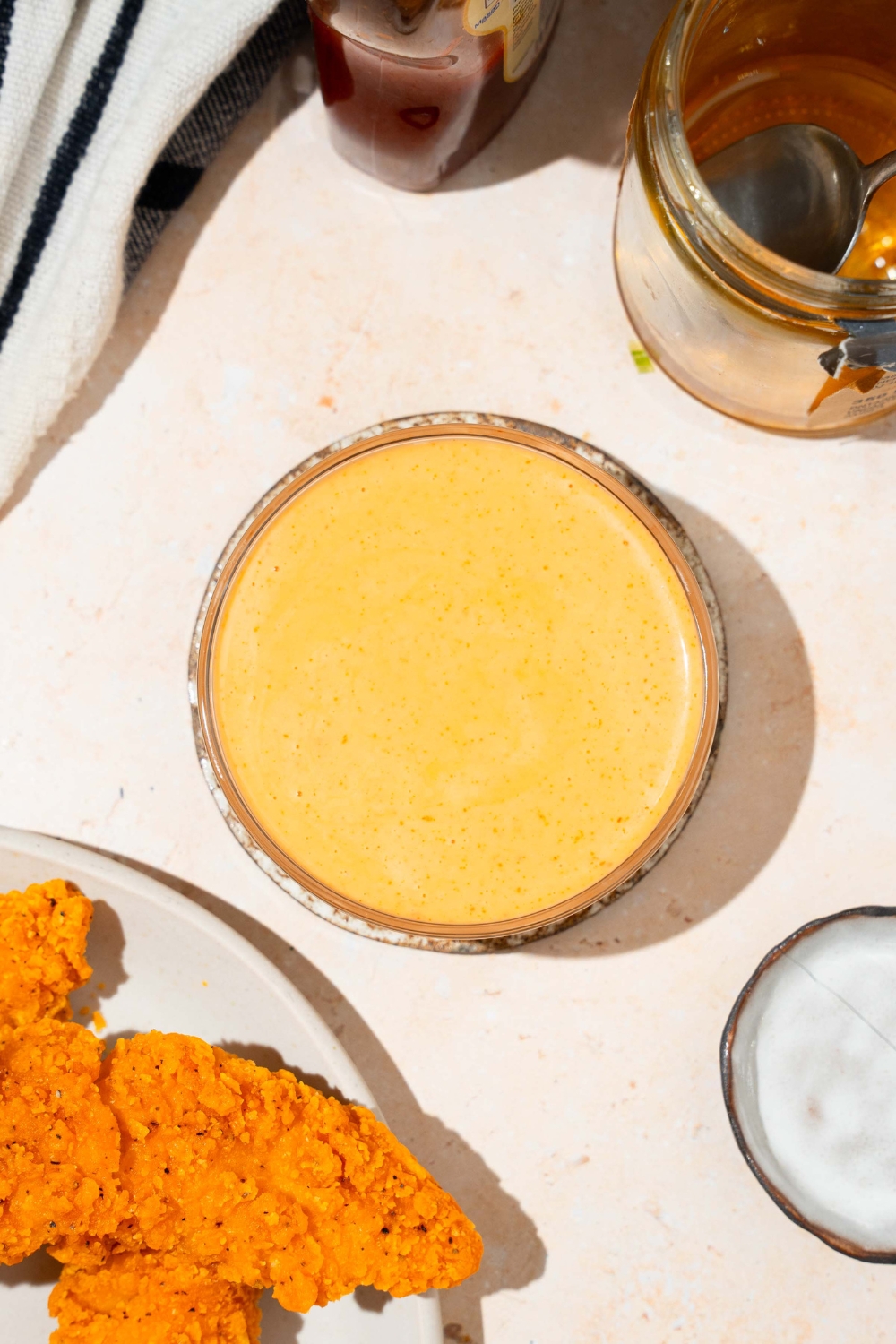 A bowl of bang bang sauce on a saucer on a tan counter with a plate of chicken tenders, jar of honey, and black striped napkin.