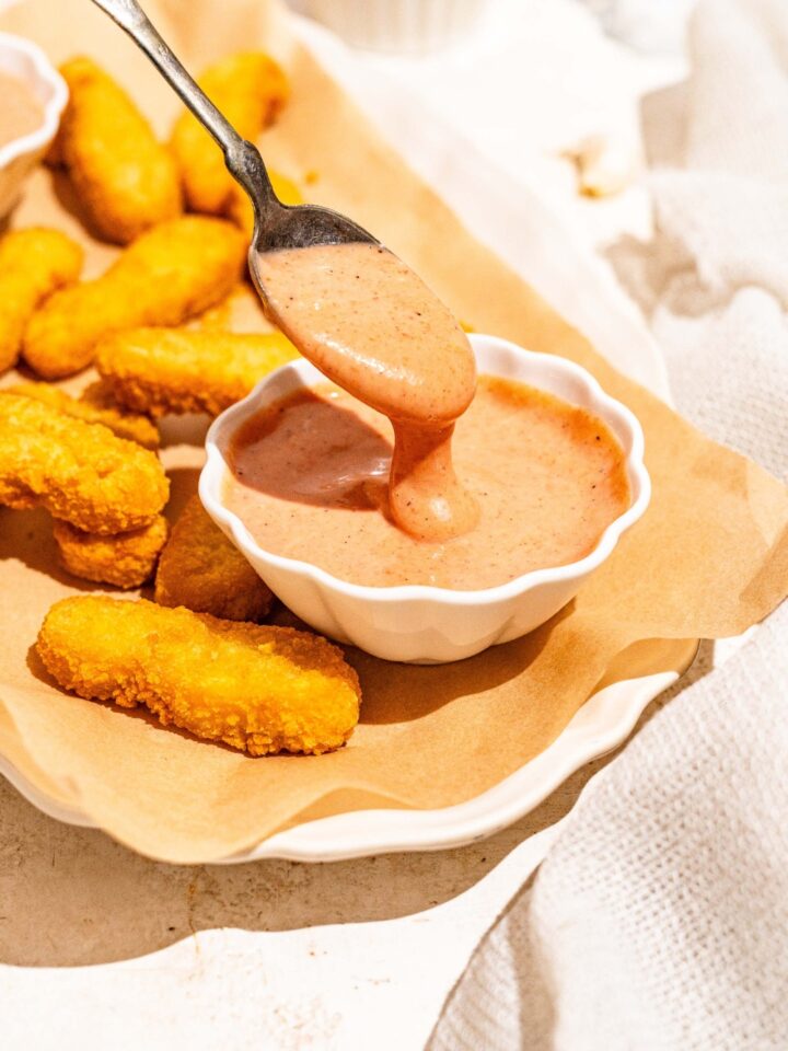 A white platter lined with parchment paper with fried fish sticks and a bowl of gochujang mayo. A spoon is dipping into the bowl. The platter is on a tan counter.