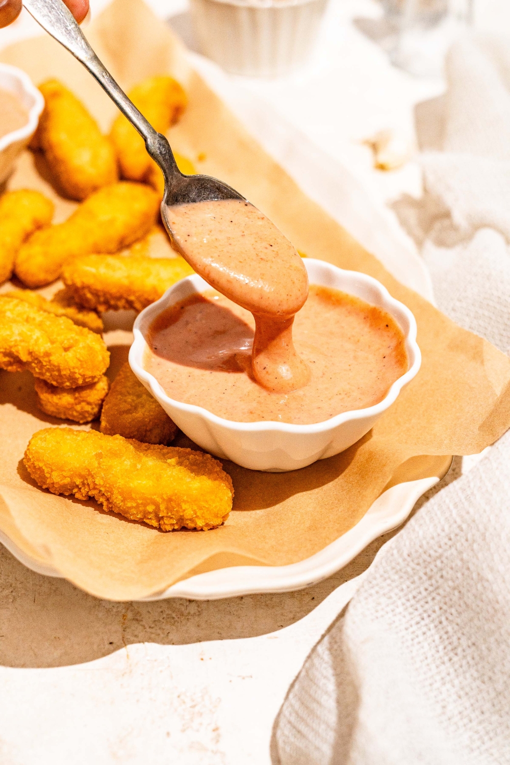 A white platter lined with parchment paper with fried fish sticks and a bowl of gochujang mayo. A spoon is dipping into the bowl. The platter is on a tan counter.