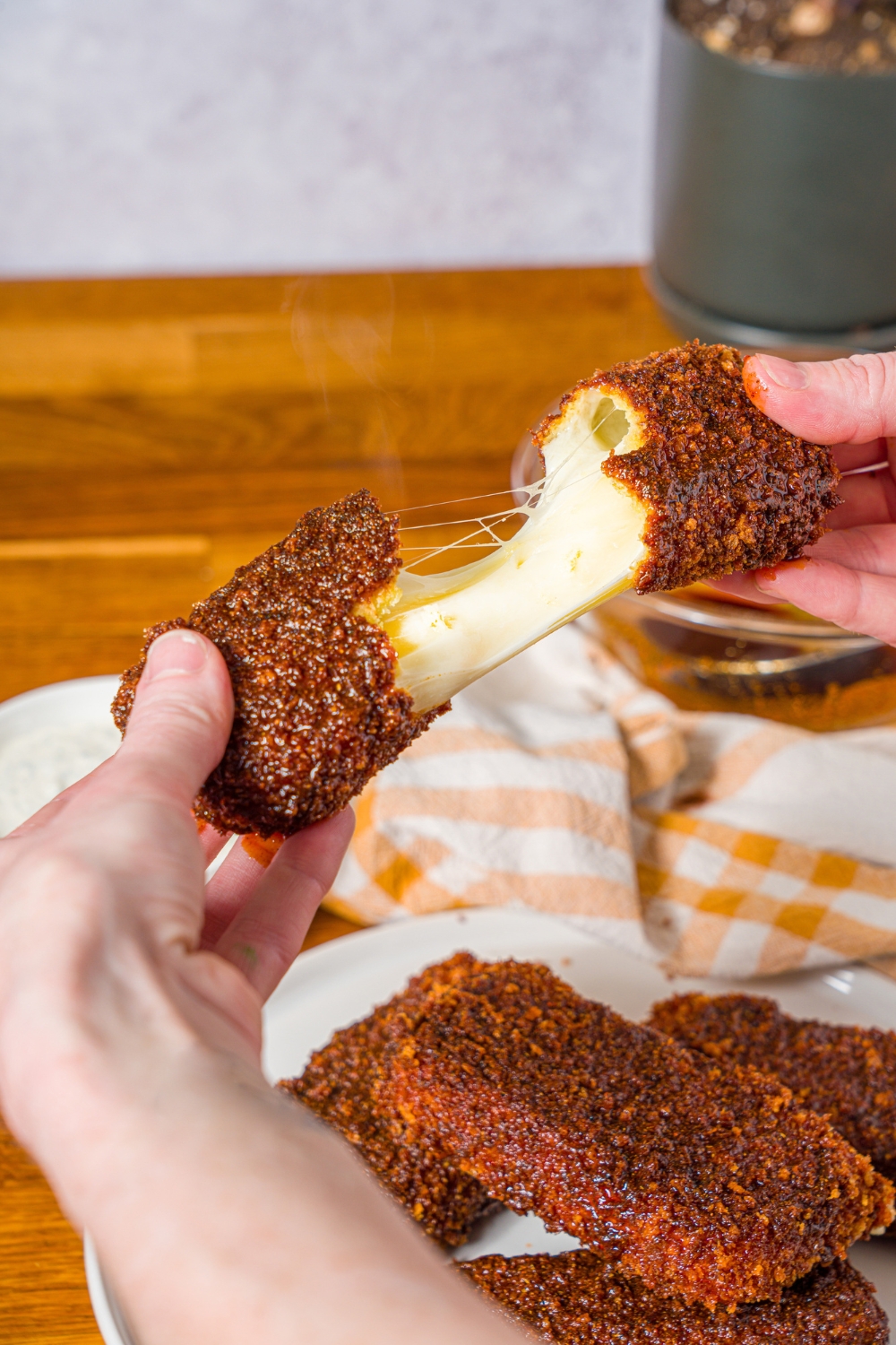 Hands pulling apart a Nashville hot mozzarella stick with a plate of mozzarella sticks. The plate is on a wooden counter with a yellow checkered napkin.