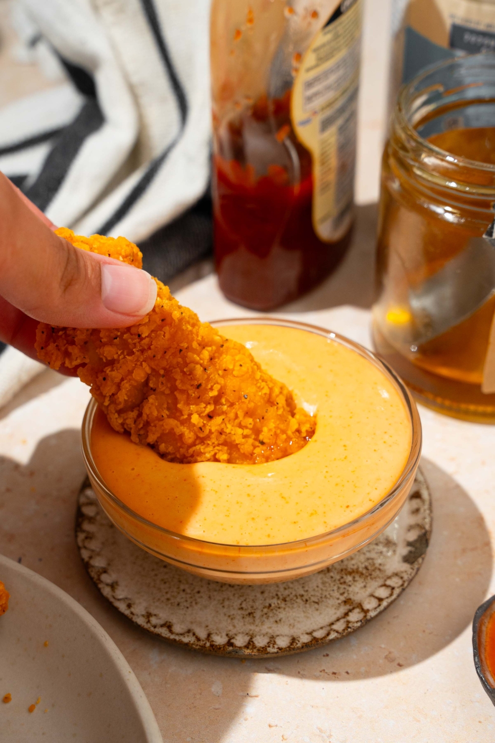 A bowl of bang bang sauce on a saucer with a chicken tender dipped in the sauce. The saucer is on a tan counter with a jar of honey, and black striped napkin.